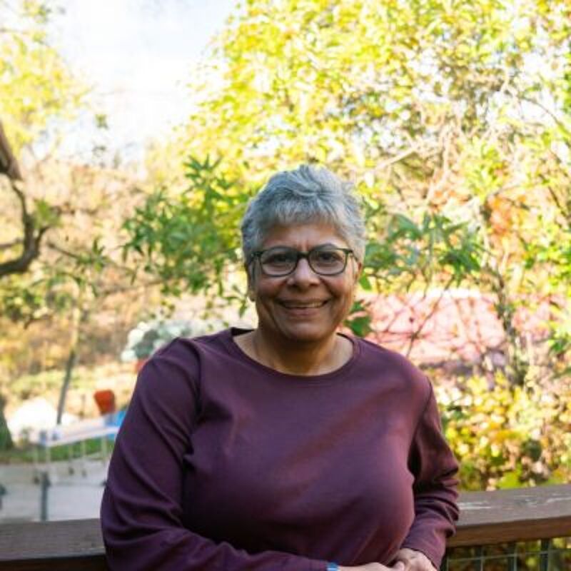 The image shows a smiling, older woman with short gray hair and glasses, wearing a long-sleeved purple shirt. She is outdoors, leaning against a wooden railing. Behind her, there's a background of green trees and foliage, suggesting a natural or park-like setting. The lighting is bright, indicating it's daytime.